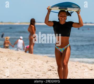 Gilgo Beach, New York, USA - 22. Juli 2023: Vorderansicht einer jungen Frau, die am Strand entlang geht, ihr Surfbrett auf dem Kopf hält und nach einem Achter lächelt Stockfoto