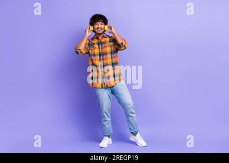 Langes Foto eines gutaussehenden Mannes mit Karohemd, Jeans, Schuhen, die hochwertige Kopfhörer berühren, isoliert auf violettem Hintergrund Stockfoto