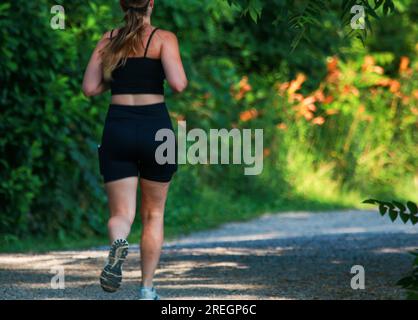 Rückansicht einer Frau, die allein im Schatten auf einem Schotterweg im Southards Pond Park an einem Sommermorgen läuft. Stockfoto