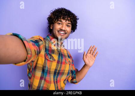 Selfie eines jungen Mannes arabische Locken Haarbekleidung Karomuster stilvolles Hemd gewellt Hand hallo Geste positive Stimmung isoliert auf violettem Hintergrund Stockfoto