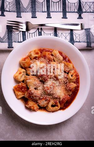 Gekochter italienischer Tortellini in roter Tomatensoße mit Käse in weißer Schüssel auf Marmoroberfläche mit grafischer Serviette Stockfoto