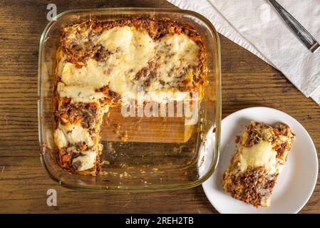 Gebackene Lasagne in einer Glaspfanne, serviert auf weißem Teller mit Gabel und Serviette auf Holzoberfläche Stockfoto