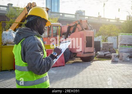 Rückansicht eines schwarzen Mannes afrikanischer ethnischer Herkunft, Ingenieur mit gelbem Sicherheitshelm, reflektierender Weste und Jacke, steht und nimmt das Konstrukt zur Kenntnis Stockfoto