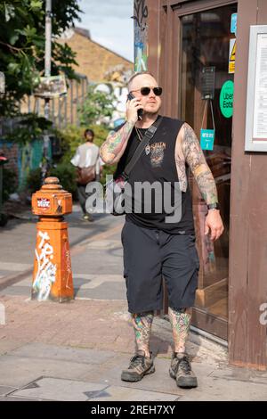 Männliche Figur mittleren Alters mit Tätowierungen, trägt schwarze Shorts und ärmelloses Hemd, und eine Sonnenbrille, die mit dem Handy spricht. Stockfoto