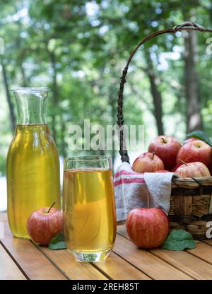 Apfelsaft im Glas und Karaffe mit frischen roten Äpfeln im Freien auf einem hölzernen Terrassentisch und Naturhintergrund Stockfoto