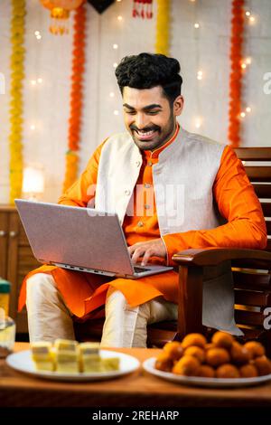 Glücklicher junger indischer Mann, der während der diwali Festival Feier im dekorierten Haus ein Notebook benutzte, vor Geschenken und Geschenken, indem er in die Kamera schaute - Konzept Stockfoto