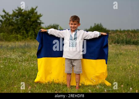 Ukrainischer Junge in besticktem Hemd-vyshyvanka, der am Sommertag auf Wiese die blau-gelbe Flagge der Ukraine demonstriert. Unabhängigkeitstag der Ukraine. Verstopfung Stockfoto