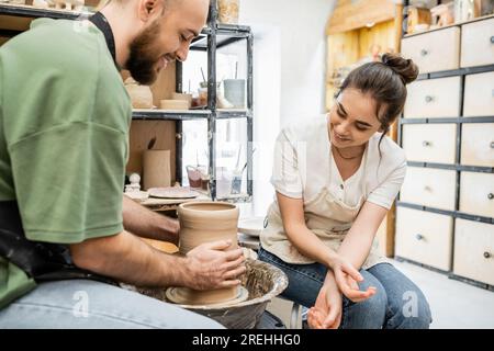 Lächelnde Handwerkerin in Schürze, die neben ihrem Freund sitzt und in der Werkstatt eine Tonvase auf einem Töpferrad macht Stockfoto