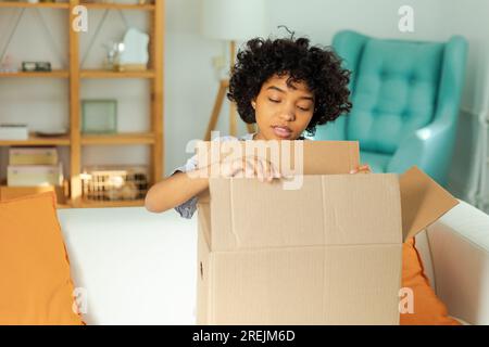 Afrikanisches Mädchen, das die Lieferung auspackt und in die Schachtel schaut. Glückliche Frau, die eine Schachtel öffnet. Eine Frau bekommt ein Paket, das sich die gelieferten Waren ansieht. Zufriedene Kunden positives Feedback, schnelle Expresslieferung Stockfoto