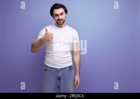 Selbstbewusster junger, brutaler brünetter Mann in einem weißen T-Shirt und Jeans zeigt Klasse mit seiner Hand Stockfoto