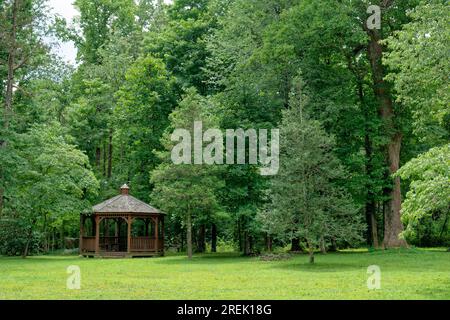 Kleiner Park mit einem großen Pavillon mit Bänken im Inneren, umgeben von einer Vielzahl von Bäumen in einer bewaldeten Umgebung und einer gemähten Rasenfläche an einem sonnigen Tag im Großen und Ganzen Stockfoto