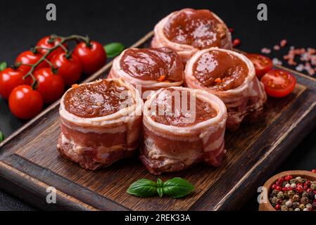 Köstliches rohes frisches Schweinefleisch oder Brötchen mit Hähnchenfleisch, eingewickelt in Speck, mit Salz, Gewürzen und Kräutern auf dunklem Betonhintergrund Stockfoto