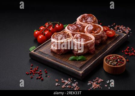 Köstliches rohes frisches Schweinefleisch oder Brötchen mit Hähnchenfleisch, eingewickelt in Speck, mit Salz, Gewürzen und Kräutern auf dunklem Betonhintergrund Stockfoto