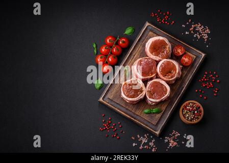 Köstliches rohes frisches Schweinefleisch oder Brötchen mit Hähnchenfleisch, eingewickelt in Speck, mit Salz, Gewürzen und Kräutern auf dunklem Betonhintergrund Stockfoto