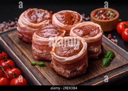 Köstliches rohes frisches Schweinefleisch oder Brötchen mit Hähnchenfleisch, eingewickelt in Speck, mit Salz, Gewürzen und Kräutern auf dunklem Betonhintergrund Stockfoto