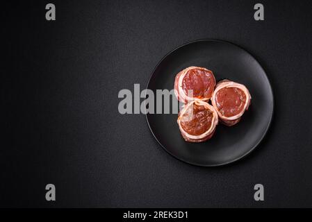 Köstliches rohes frisches Schweinefleisch oder Brötchen mit Hähnchenfleisch, eingewickelt in Speck, mit Salz, Gewürzen und Kräutern auf dunklem Betonhintergrund Stockfoto