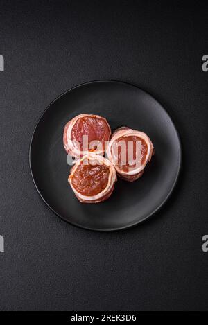 Köstliches rohes frisches Schweinefleisch oder Brötchen mit Hähnchenfleisch, eingewickelt in Speck, mit Salz, Gewürzen und Kräutern auf dunklem Betonhintergrund Stockfoto