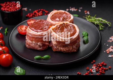 Köstliches rohes frisches Schweinefleisch oder Brötchen mit Hähnchenfleisch, eingewickelt in Speck, mit Salz, Gewürzen und Kräutern auf dunklem Betonhintergrund Stockfoto