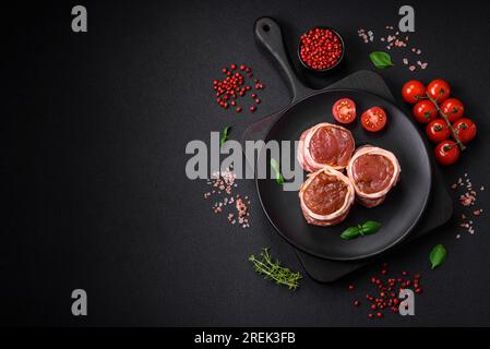 Köstliches rohes frisches Schweinefleisch oder Brötchen mit Hähnchenfleisch, eingewickelt in Speck, mit Salz, Gewürzen und Kräutern auf dunklem Betonhintergrund Stockfoto