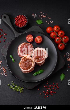 Köstliches rohes frisches Schweinefleisch oder Brötchen mit Hähnchenfleisch, eingewickelt in Speck, mit Salz, Gewürzen und Kräutern auf dunklem Betonhintergrund Stockfoto