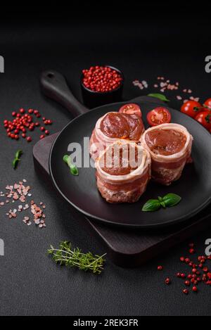 Köstliches rohes frisches Schweinefleisch oder Brötchen mit Hähnchenfleisch, eingewickelt in Speck, mit Salz, Gewürzen und Kräutern auf dunklem Betonhintergrund Stockfoto