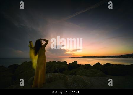 Frau in gelbem Kleid und Umhang, die einen herrlichen Sonnenaufgang an der französischen baskischen Küste II beobachtet Stockfoto