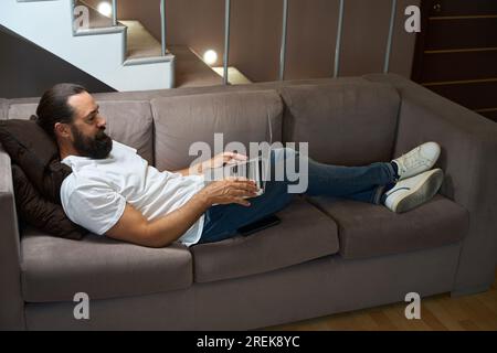 Ein Mann mittleren Alters liegt auf einem Sofa mit einem Laptop Stockfoto