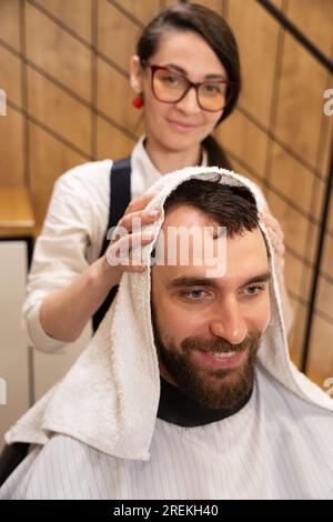 Der süße Friseur trocknet die Haare des Kunden mit einem weichen Handtuch Stockfoto