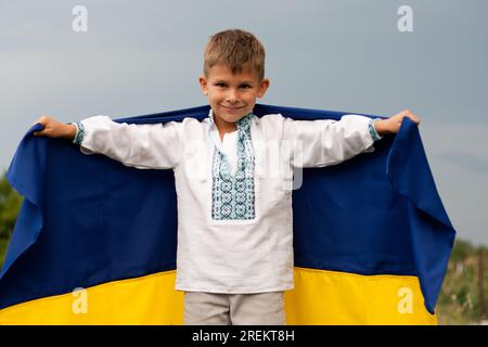 Glücklicher ukrainischer Junge in besticktem Hemd - vyshyvanka mit blau-gelber Flagge der Ukraine, gegen den Himmel. Unabhängigkeitstag der Ukraine. Verfassungstag. Da Stockfoto