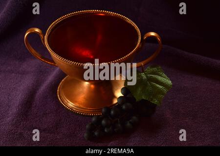 Goldfarbener Kelch mit roter Flüssigkeit und Trauben daneben auf einem violetten Tuch Stockfoto