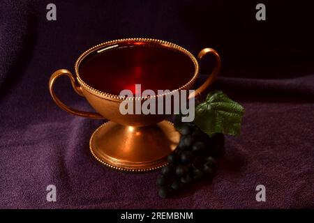 Goldfarbener Kelch mit roter Flüssigkeit und Trauben daneben auf einem violetten Tuch Stockfoto