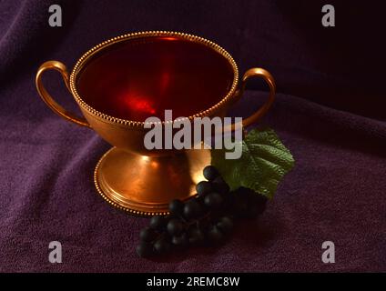 Goldfarbener Kelch mit roter Flüssigkeit und Trauben daneben auf einem violetten Tuch Stockfoto