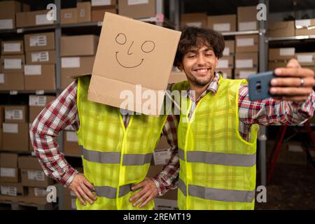 Glückliches Lagerteam mit Box auf dem Kopf, das ein Selfie in Arbeitskleidung macht. Multiethnische Logistikmitarbeiter arbeiten in einem großen Distrikt zusammen Stockfoto