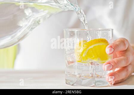 Weibliche Hände gießen Wasser aus der Karaffe in einen Glasbecher mit Zitrone und Eis Stockfoto