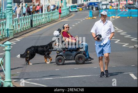 Mann in einem elektrischen Mobilitätsroller mit einem Hund an einem Bleistift, überquert eine Straße in England, Großbritannien. Stockfoto