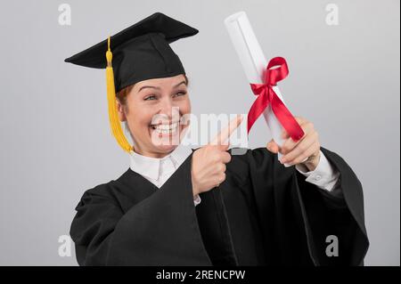 Glückliche Frau im Graduiertenkleid, die mit dem Finger auf ein Diplom zeigt. Stockfoto