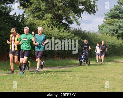 Kesgrave, Suffolk - 29. Juli 2023 : warmer Sommer am Samstagmorgen Parkrun im Millennium Field. Daumen hoch von einem lächelnden Läufer. Der Buggy kommt von hinten. Stockfoto