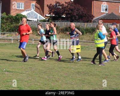 Kesgrave, Suffolk - 29. Juli 2023 : warmer Sommer am Samstagmorgen Parkrun im Millennium Field. Die Gruppe fährt am Marshal an der Ecke High 5 vorbei. Stockfoto