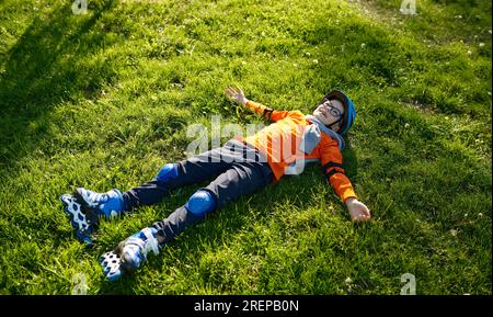 Fröhlicher, müder Junge mit Rollschuhen und Helm auf grünem Gras Stockfoto