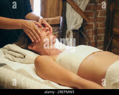 Nahaufnahme der Hände eines Chiropraktikers oder Masseurs, der eine entspannende Gesichtsmassage für eine liegende junge, fit Frau in der Wellness-Spa-Hütte durchführt. Stockfoto