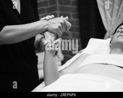 Nicht wiedererkennbare weibliche Masseusenhände, die Finger- und Handmassagen an eine junge, weiße Kundin, die auf einem Massagetisch im Spa-Salon liegt, machen Stockfoto