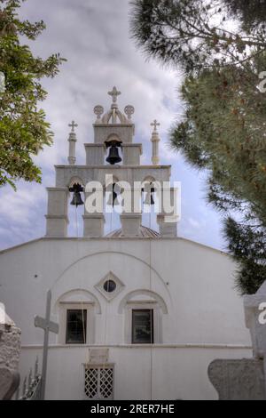 Glockenturm einer griechisch-orthodoxen Kirche in einem Dorf auf der Insel Naxos, Griechenland. Umrahmt von Bäumen vor einem leicht bewölkten Himmel. Stockfoto