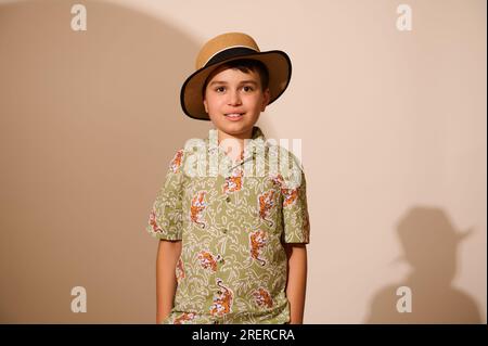 Ein süßer Teenager mit Strohhut und einem stilvollen, gemusterten Sommershirt, der seine Hände in die Tasche steckt, lächelt in die Kamera, isoliert auf Studiohintergrund Stockfoto