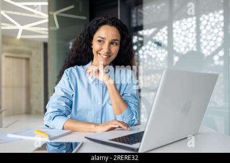 Junge, schöne, denkende Frau, die lächelt und aus dem Fenster schaut, Geschäftsfrau, die am Arbeitsplatz lächelt, zufrieden mit den erzielten Ergebnissen, lateinamerikanische Frau, die bei der Arbeit ein Notebook verwendet. Stockfoto