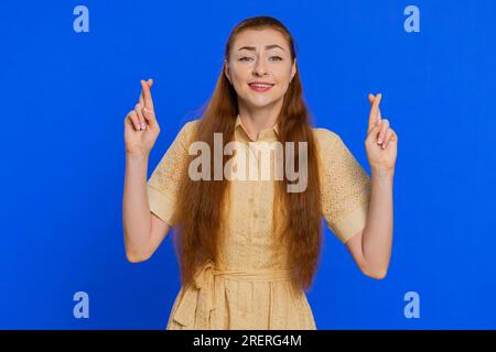 Aufgeregte, schockierte Frau drückte die Daumen, als sie in die Kamera sah und um Glücksnachrichten bat, wünschte gute Prüfungsergebnisse, träumte vom Sieg im Lotto-Jackpot. Mädchen isoliert auf blauem Studiohintergrund Stockfoto