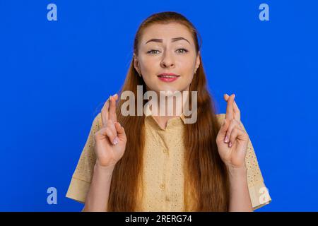 Aufgeregte, schockierte Frau drückte die Daumen, als sie in die Kamera sah und um Glücksnachrichten bat, wünschte gute Prüfungsergebnisse, träumte vom Sieg im Lotto-Jackpot. Mädchen isoliert auf blauem Studiohintergrund Stockfoto