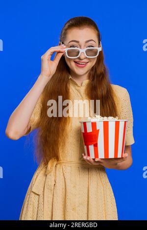 Aufgeregte junge Erwachsene Frau mit 3D-Brille, die Popcorn isst und interessante fernsehserien, Sportspiele, Online-Filme in sozialen Medien ansieht. Rothaarige Frau isoliert auf blauem Studiohintergrund. Vertikal Stockfoto