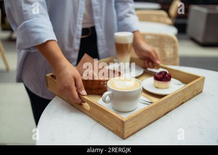 Eine gut lächelnde Kellnerin in einem Restaurant hielt ein Tablett mit einem Latte Kaffee, Cappuccino und Beerenkuchen in einem Restaurant Stockfoto
