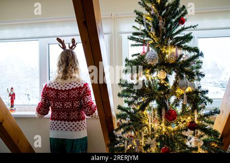 Nicht wiedererkennbare Erwachsene Frau in rotem weihnachtlichem Pullover, die aus dem Fenster schaut und auf Weihnachtsfeiertage neben dem Weihnachtsbaum wartet. Stockfoto