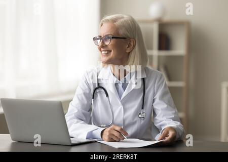 Glückliche, wohlüberlegte, reife Ärztin, die am Arbeitsplatz sitzt Stockfoto
